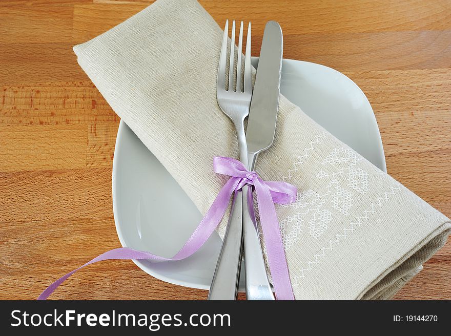White plate, fork and knife, tied with purple ribbon, with a linen cloth on a wooden table. White plate, fork and knife, tied with purple ribbon, with a linen cloth on a wooden table