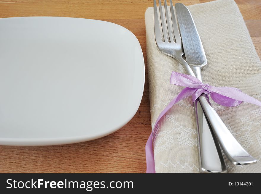 White plate, fork and knife, tied with purple ribbon, with a linen cloth on a wooden table. White plate, fork and knife, tied with purple ribbon, with a linen cloth on a wooden table