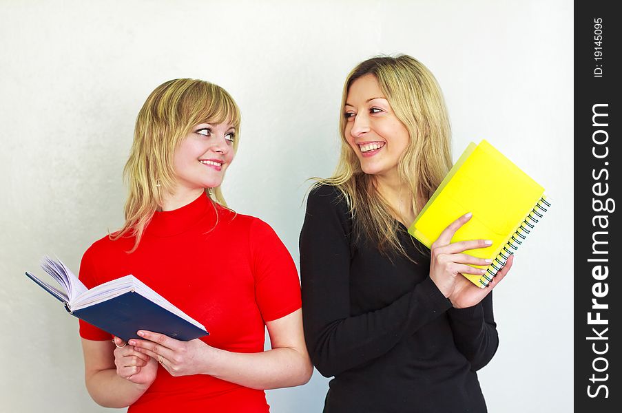 Two beautiful young girl with long blond hair are holding a book and notebook, look at each other, smiling. Two beautiful young girl with long blond hair are holding a book and notebook, look at each other, smiling