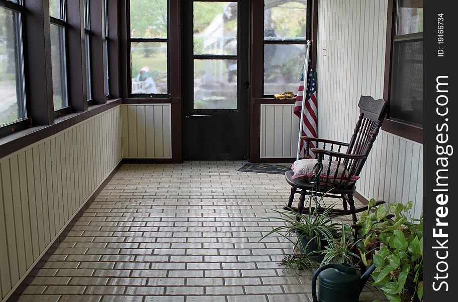 Enclosed, tiled, porch with potted plants, a watering can, an antique rocker and an American Flag. Enclosed, tiled, porch with potted plants, a watering can, an antique rocker and an American Flag.