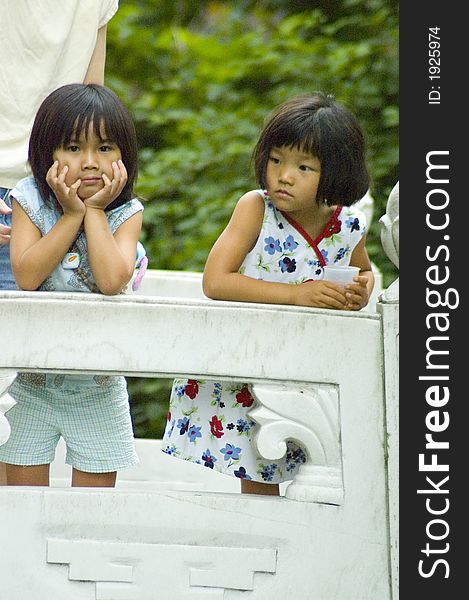 Two cute young Asian girls lean on the rail of a stone bridge, one thoughtfully holding her face in her hands and looking at the camera. Two cute young Asian girls lean on the rail of a stone bridge, one thoughtfully holding her face in her hands and looking at the camera.