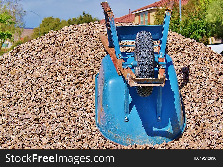 Wheelbarrow on Brown Gravel Pile in Front of Houses and Sky. Wheelbarrow on Brown Gravel Pile in Front of Houses and Sky