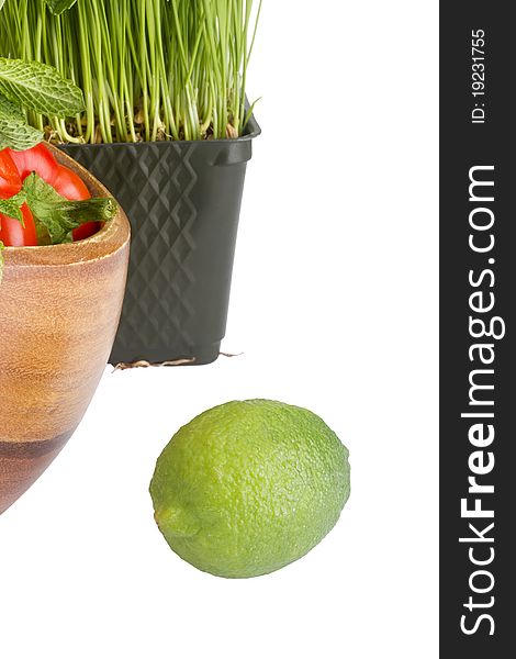 Green lime isolated on a white background next to wheatgrass and a wooden plate with red bell peppers.