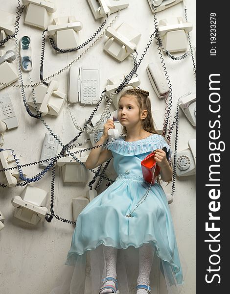 Image of a beautiful seriously little girl sitting on the white cube and holding a phone surrounded by many phones on the wall. Image of a beautiful seriously little girl sitting on the white cube and holding a phone surrounded by many phones on the wall