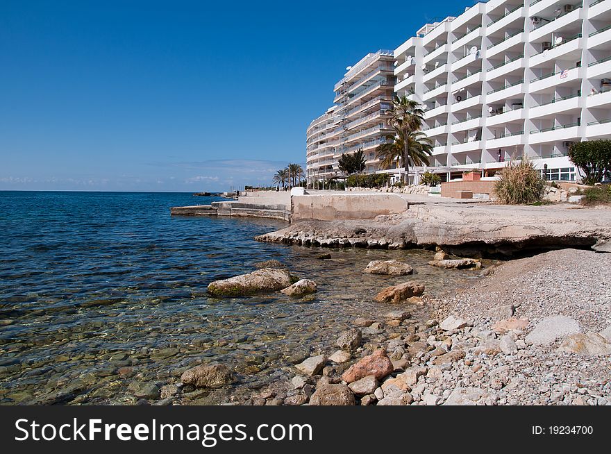 Image shows coast of Santa Ponsa, Majorca, Spain. Blue sea on the right, hotels and some stones on the right, and blue sky above. Image shows coast of Santa Ponsa, Majorca, Spain. Blue sea on the right, hotels and some stones on the right, and blue sky above.