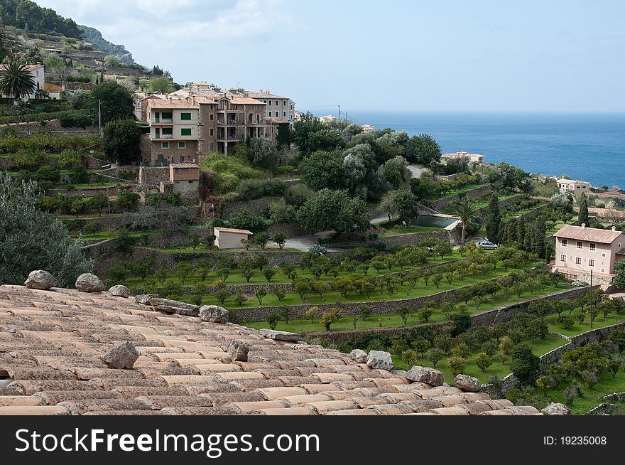 Image shows coast of Banyalbufar, Majorca, Spain. In front an old roof, in the middle some green trees, stone walls and small houses in front of blue sky and blue sea.