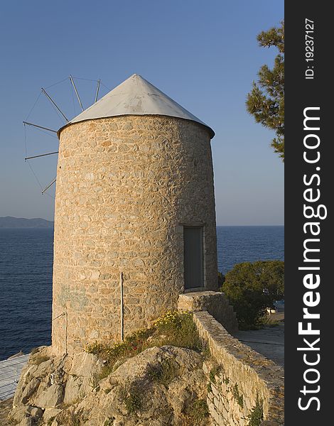 Old mill built of stone lit with late afternoon light surrounded by spring flowers and set in front of Aegean Sea on Greek island Hydra. Old mill built of stone lit with late afternoon light surrounded by spring flowers and set in front of Aegean Sea on Greek island Hydra.