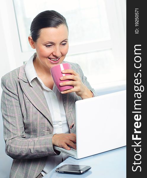 Attractive smiling young business woman with cup using laptop at work desk