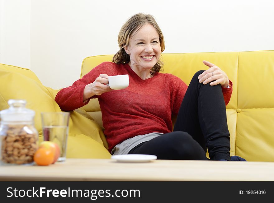 Woman in a red top shares a laugh while she relaxes on a sofa with a hot drink. Woman in a red top shares a laugh while she relaxes on a sofa with a hot drink