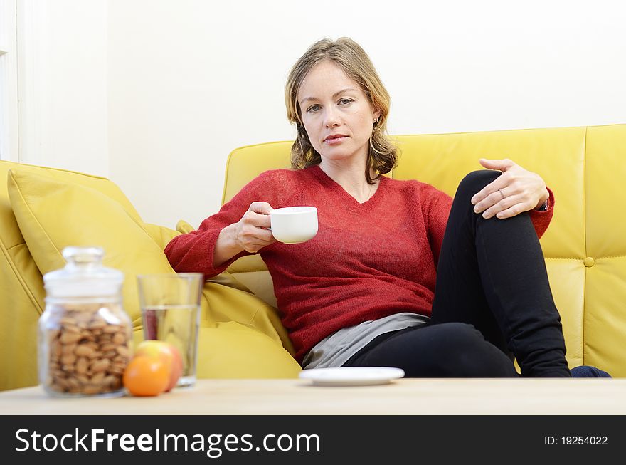 Woman sitting on a yellow leather sofa looking serious and holding a hot drink. Woman sitting on a yellow leather sofa looking serious and holding a hot drink