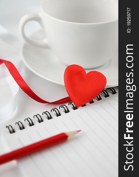 White tea cup with note-pad, pencil and red heart.  Soft focus. White tea cup with note-pad, pencil and red heart.  Soft focus