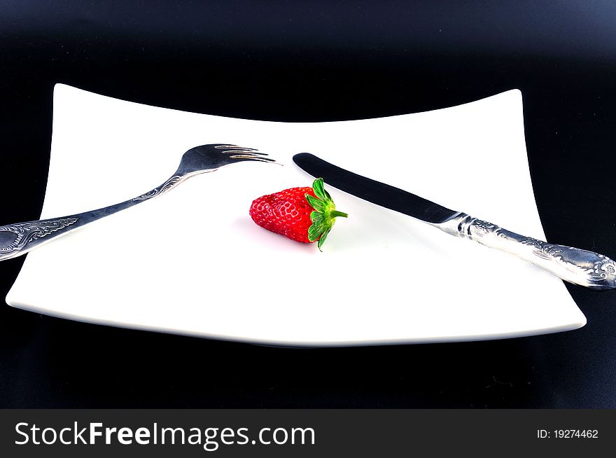 Knife and fork are on a white plate next to a strawberry. Knife and fork are on a white plate next to a strawberry