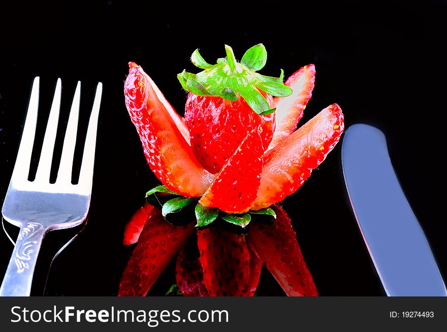 Knife and fork are close to the cut strawberries against a black background. Knife and fork are close to the cut strawberries against a black background