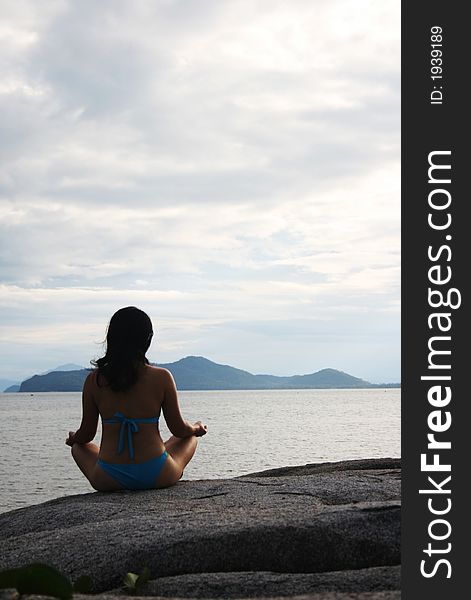 A girl in blue bikini sitted cross-legged on a rock by the beach, meditating. A girl in blue bikini sitted cross-legged on a rock by the beach, meditating.