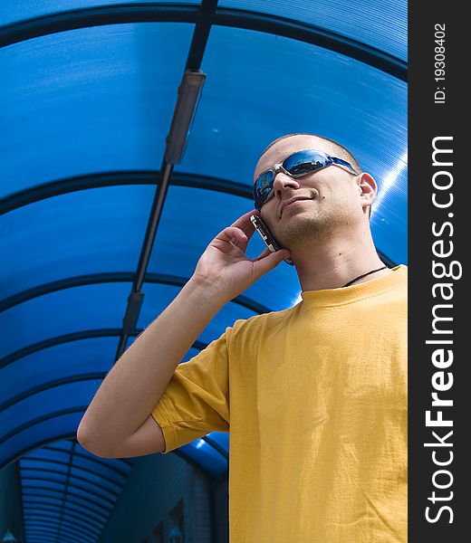 Man talking on the phone, wearing a yellow t-shirt and blue sun glasses, against blue background. Man talking on the phone, wearing a yellow t-shirt and blue sun glasses, against blue background