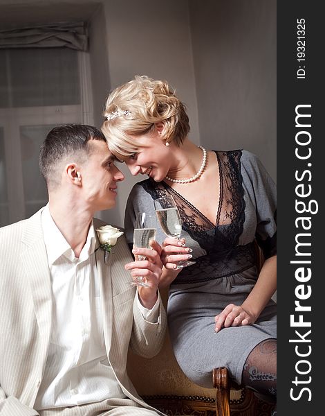 Happy bride in gray dress and groom wearing white suite in bedroom interior with glasses. Happy bride in gray dress and groom wearing white suite in bedroom interior with glasses