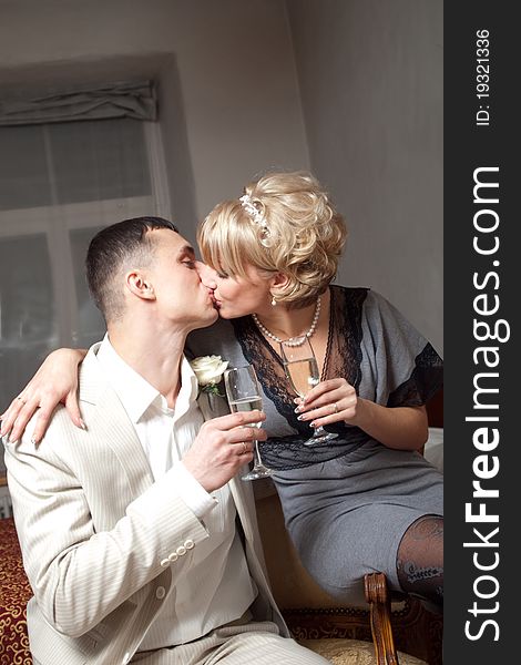 Happy bride in gray dress and groom wearing white suite are kissing in bedroom interior with glasses. Happy bride in gray dress and groom wearing white suite are kissing in bedroom interior with glasses