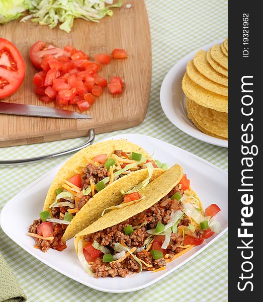 Two tacos with lettuce, tomatoes, cheese on a plate with ingredients in background