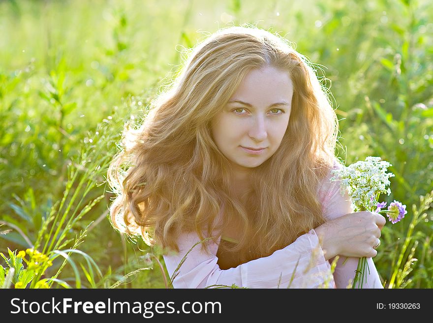 A young girl with blond long hair sitting in a field with a bouquet of flowers in the rays of evening sun