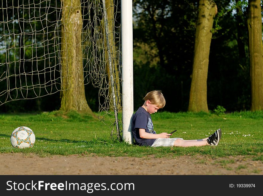 Boy playing a computer game leaning against a goal post on a football field. Boy playing a computer game leaning against a goal post on a football field