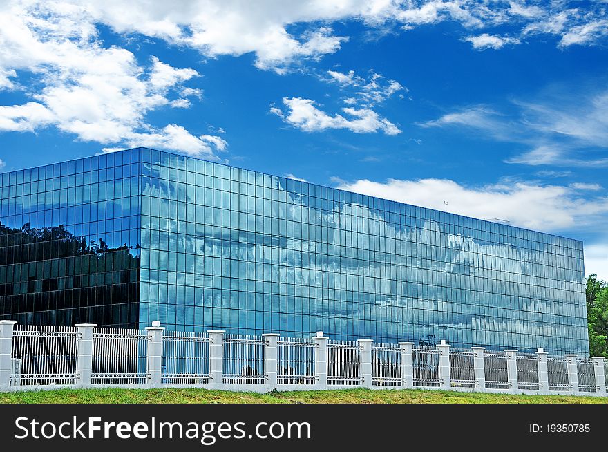 A simple modern stylish rectangular building with blue tint reflective glass all around exterior, decorated stainless steel compound fence with concrete post also constructed, blue sky with clouds background. A simple modern stylish rectangular building with blue tint reflective glass all around exterior, decorated stainless steel compound fence with concrete post also constructed, blue sky with clouds background