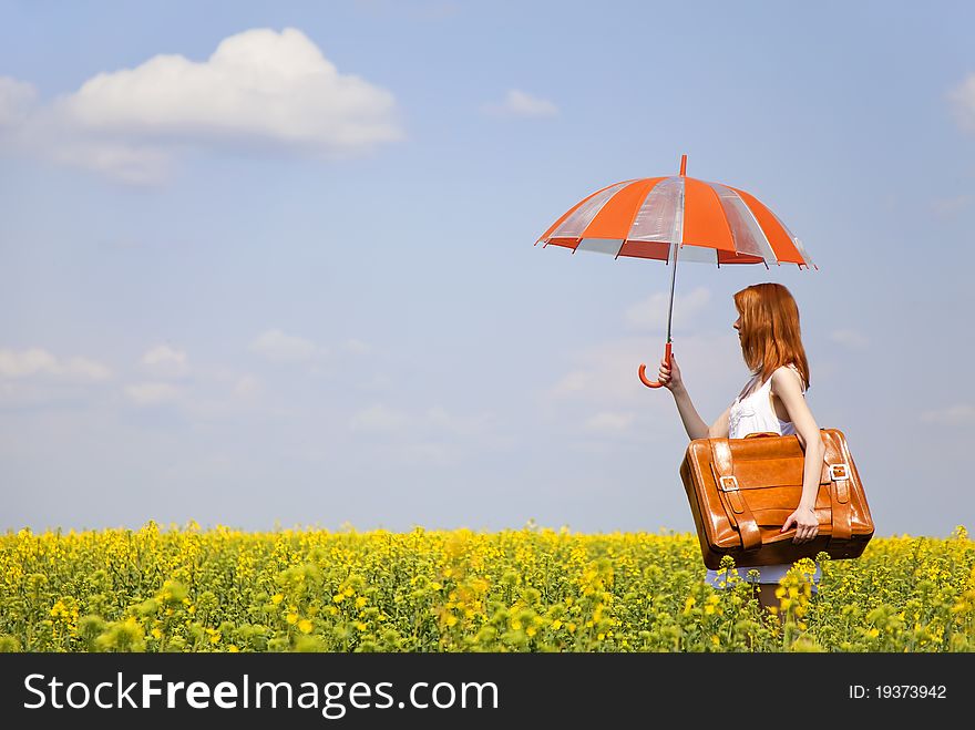 Redhead enchantress with umbrella and suitcase at spring rapeseed field.