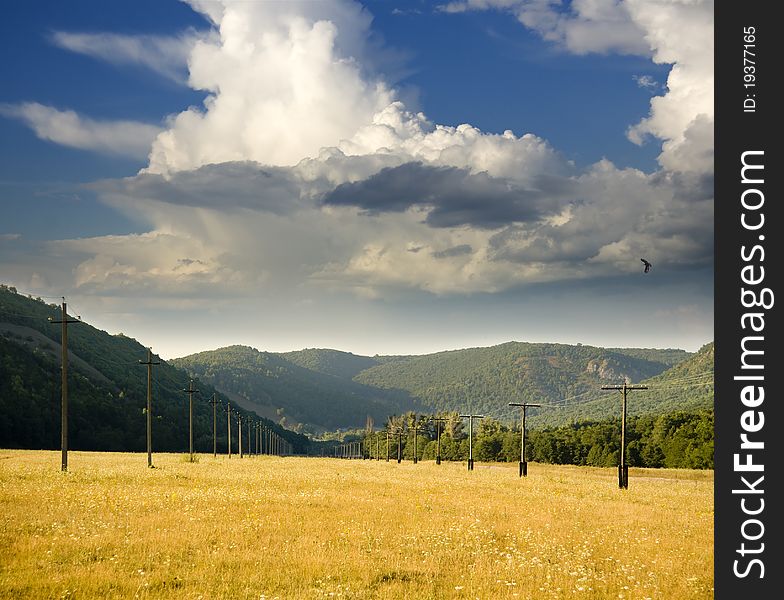 Power line is located in a mountain valley in the pasture with dry grass on a background of white large clouds. Power line is located in a mountain valley in the pasture with dry grass on a background of white large clouds.