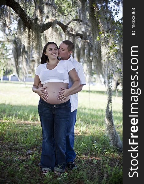 A young man kissing the cheek of a pregnant woman outside under a tree. A young man kissing the cheek of a pregnant woman outside under a tree.