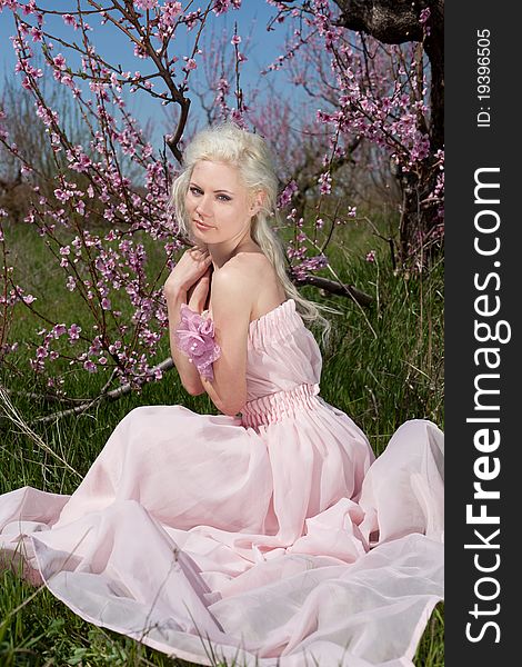 Outdoor shot of Young pretty blond woman wearing pink dress sitting on the grass in blooming garden. Outdoor shot of Young pretty blond woman wearing pink dress sitting on the grass in blooming garden