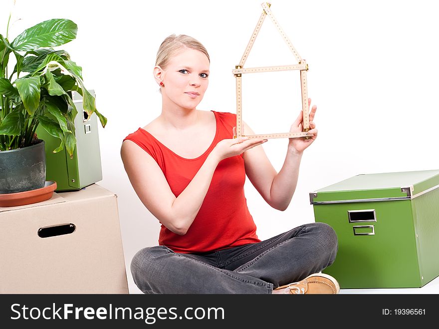 Young woman with a folding rule as a house symbol sits between move cardboards and a plant. Young woman with a folding rule as a house symbol sits between move cardboards and a plant