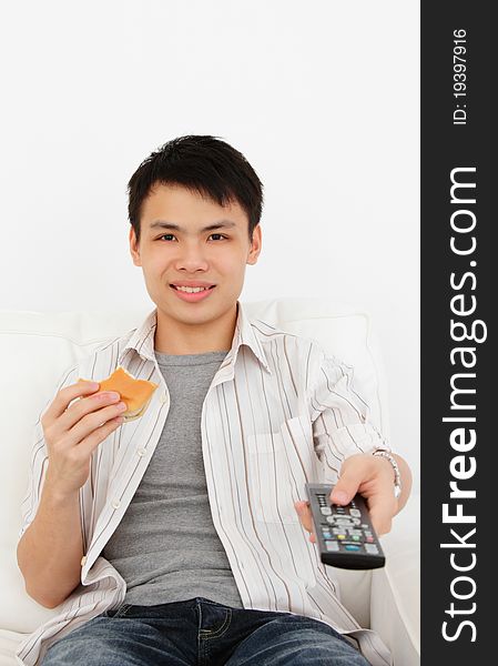 A young Asian man with a TV remote and burger on a sofa against a white background. A young Asian man with a TV remote and burger on a sofa against a white background