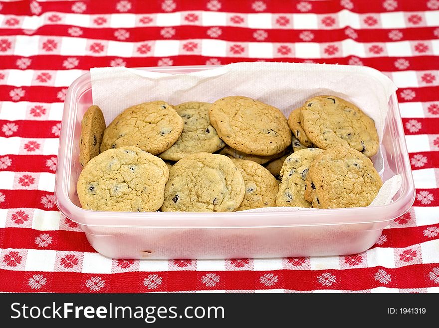 A container of homemade chocolate chip cookies on gingham tablecloth. A container of homemade chocolate chip cookies on gingham tablecloth.