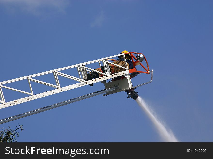 A firefighter atop a ladder sprays water onto a burning building against a clear blue sky.
