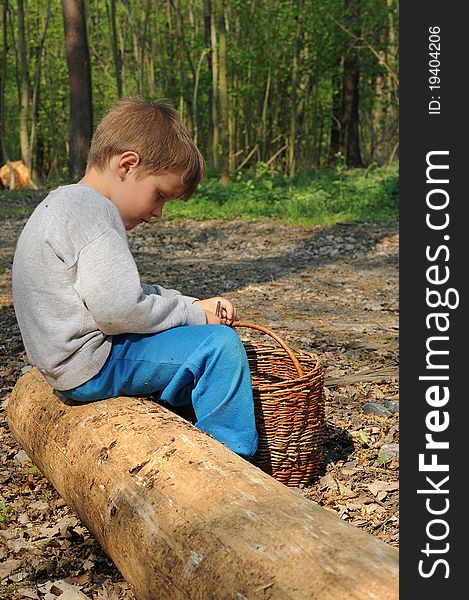 Young boy with a wicker basket sitting and having rest on a trunk in the middle of a forest. Young boy with a wicker basket sitting and having rest on a trunk in the middle of a forest