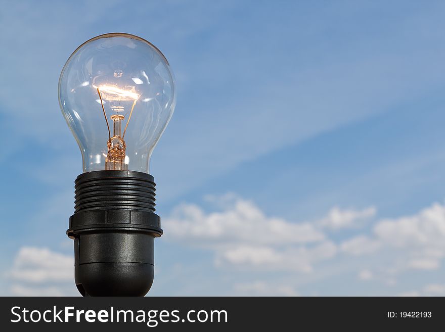 Clear electric light bulb glowing against blue sky and puffy cloud background