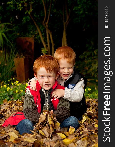 Two happy and smiling brothers or sons are sitting and hugging in a pile of colorful yellow and brown autumn / fall leaves in a garden or park setting. Two happy and smiling brothers or sons are sitting and hugging in a pile of colorful yellow and brown autumn / fall leaves in a garden or park setting