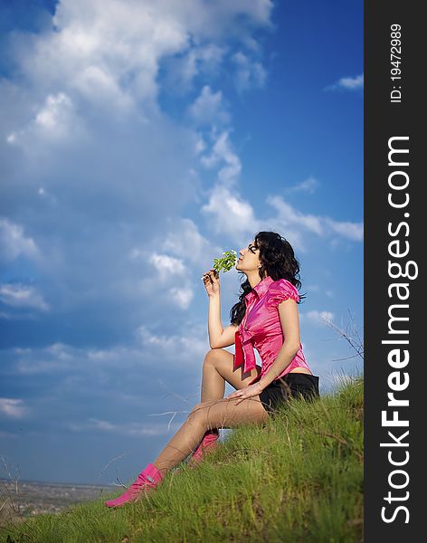Beautiful girl in pink blouse and short skirt sitting at the top of the hill on a background of blue sky and sniffs a flower. Beautiful girl in pink blouse and short skirt sitting at the top of the hill on a background of blue sky and sniffs a flower.