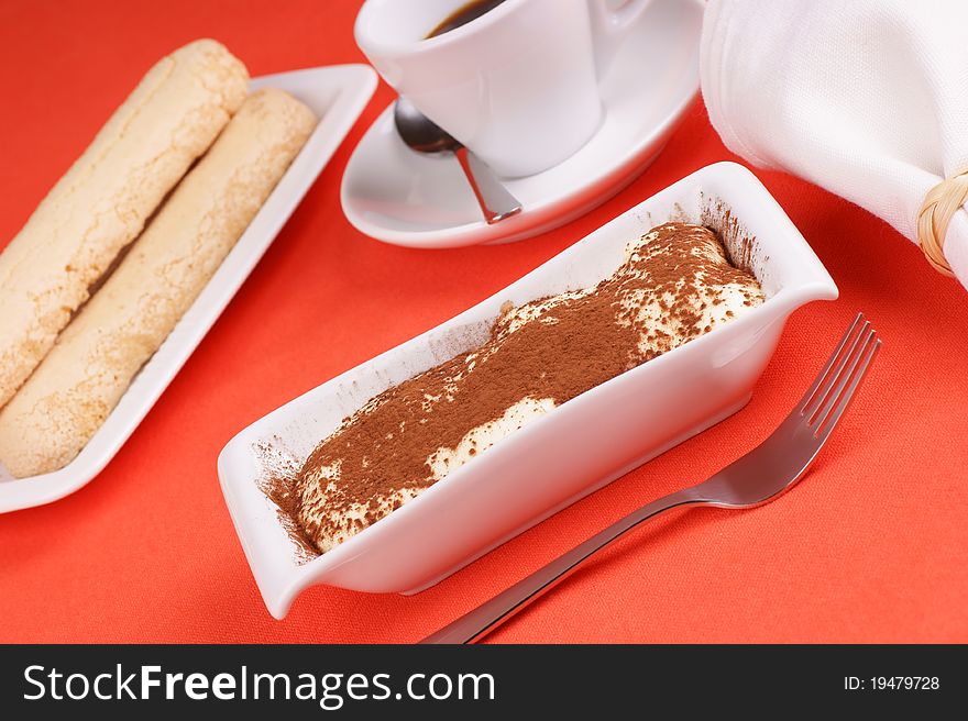 Tiramisu' cake served on a white plate. Savoiardi and cup of coffee out of focus in the background