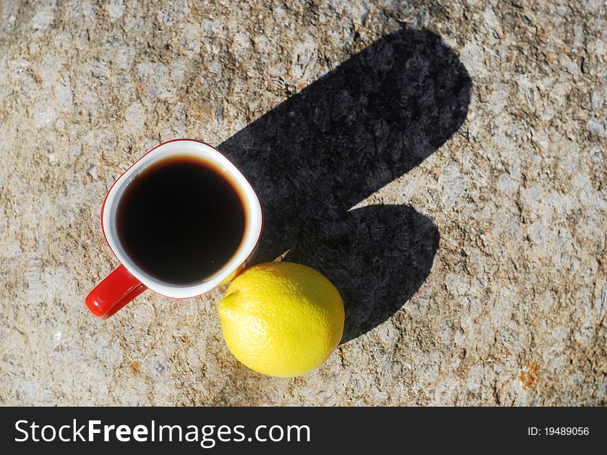 Red cup of black tea and a whole lemon fruit close-up on sunny day with long shadows outdoors. Stone background, view from above. Red cup of black tea and a whole lemon fruit close-up on sunny day with long shadows outdoors. Stone background, view from above