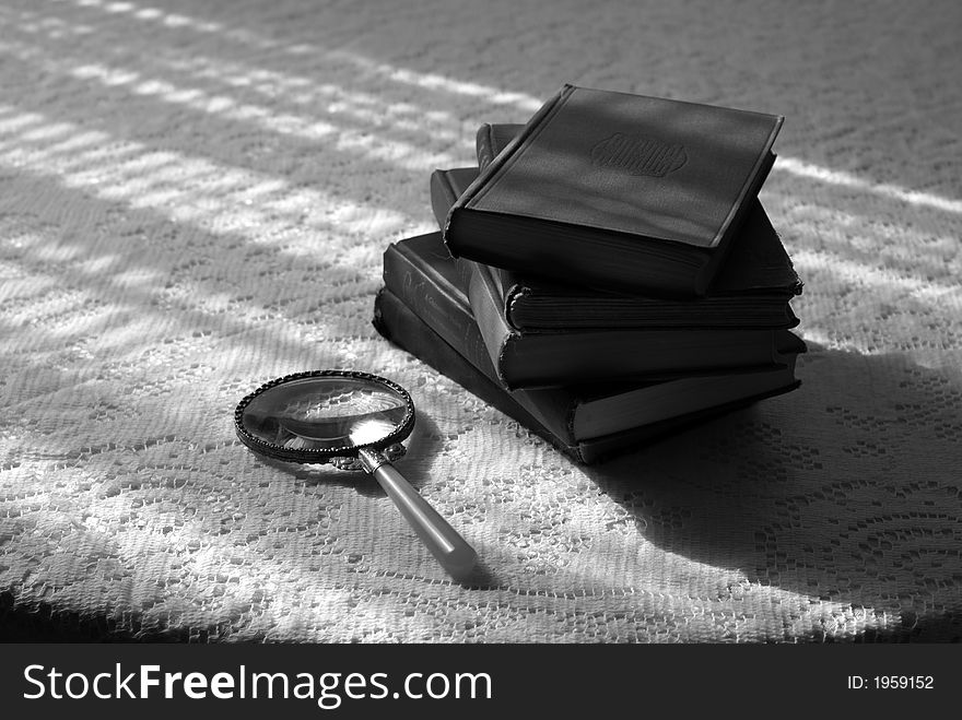 Stack of old books with reading glass on sunlit laced tabletop, black and white. Stack of old books with reading glass on sunlit laced tabletop, black and white