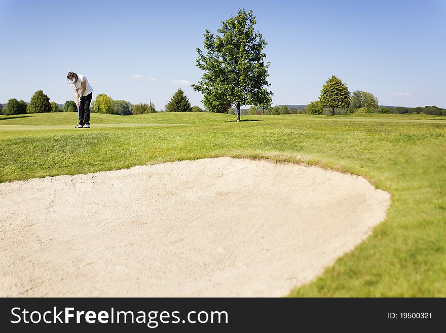 Active senior female golf player putting golf ball on green on beautiful golf course with sand bunker in foreground. Active senior female golf player putting golf ball on green on beautiful golf course with sand bunker in foreground.