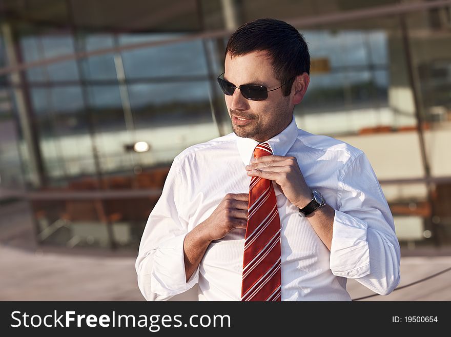 Close up of confident businessman in white shirt adjusting his orange tie, standing outdoors in front of glass office building. Close up of confident businessman in white shirt adjusting his orange tie, standing outdoors in front of glass office building.