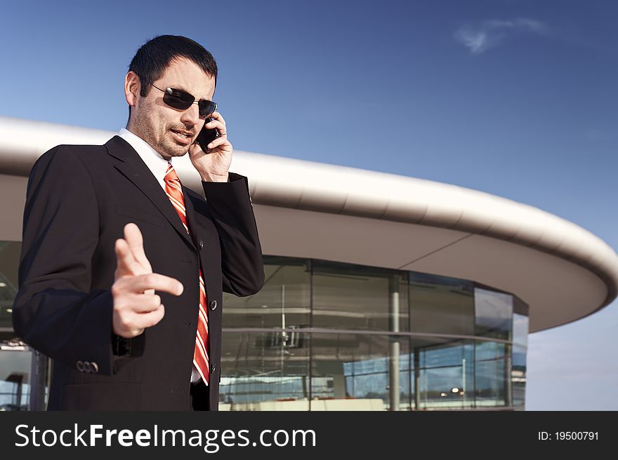 Young businessman in black suit and sunglasses being busy on cell phone with office building and blue sky in background. Young businessman in black suit and sunglasses being busy on cell phone with office building and blue sky in background.