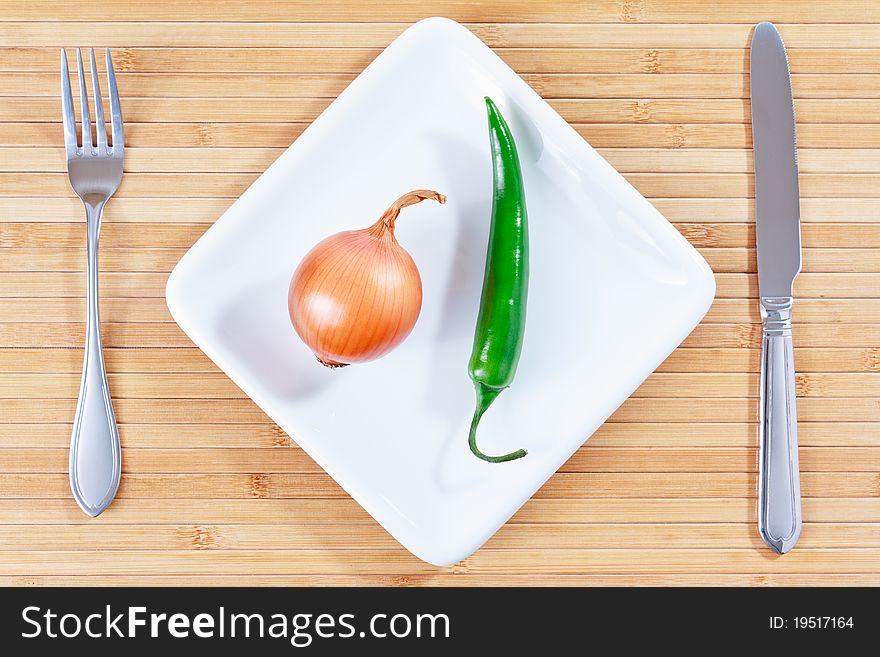 Green chili pepper and onion on a white plate and bamboo placemat with fork and knife