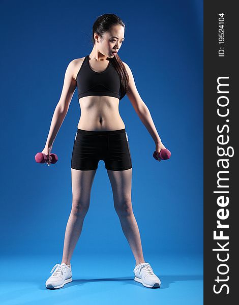 Beautiful young Asian Chinese girl working out with hand weights, wearing black sports bra and shorts, standing in blue studio. Beautiful young Asian Chinese girl working out with hand weights, wearing black sports bra and shorts, standing in blue studio.