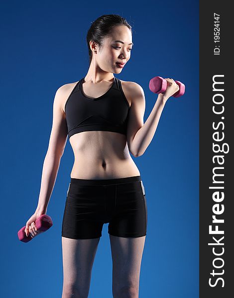 Beautiful young oriental woman working out with fitness hand weights, wearing black sports bra and shorts, standing in blue studio. Beautiful young oriental woman working out with fitness hand weights, wearing black sports bra and shorts, standing in blue studio.