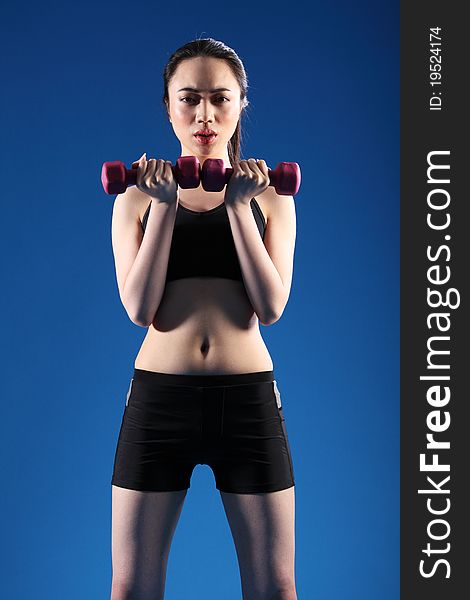Beautiful young Asian Chinese girl working out with hand weights, wearing black sports bra and shorts, standing in blue studio. Beautiful young Asian Chinese girl working out with hand weights, wearing black sports bra and shorts, standing in blue studio.