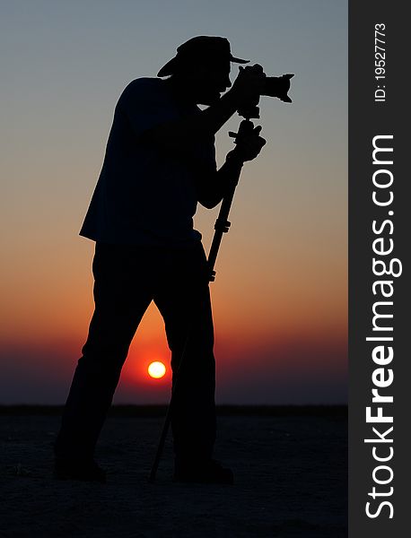Silhouette of a young man in a hat taking pictures at sunset using a tripod. Silhouette of a young man in a hat taking pictures at sunset using a tripod