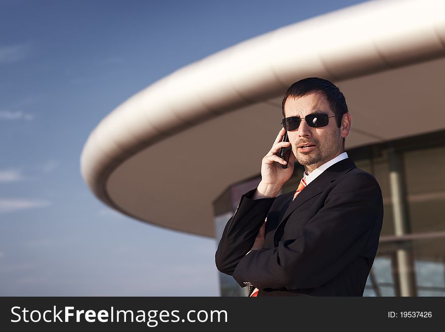 Close up of young cool business person in black suit and sunglasses being busy on cell phone with office building and blue sky in background. Close up of young cool business person in black suit and sunglasses being busy on cell phone with office building and blue sky in background.