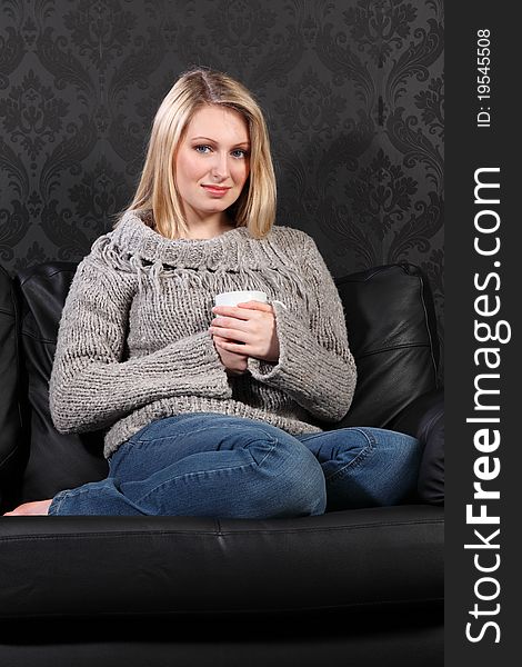 Quiet time for beautiful young blonde woman sitting with feet up on black leather sofa at home drinking coffee, wearing casual grey knitted sweater, blue jeans and just relaxing. Quiet time for beautiful young blonde woman sitting with feet up on black leather sofa at home drinking coffee, wearing casual grey knitted sweater, blue jeans and just relaxing.